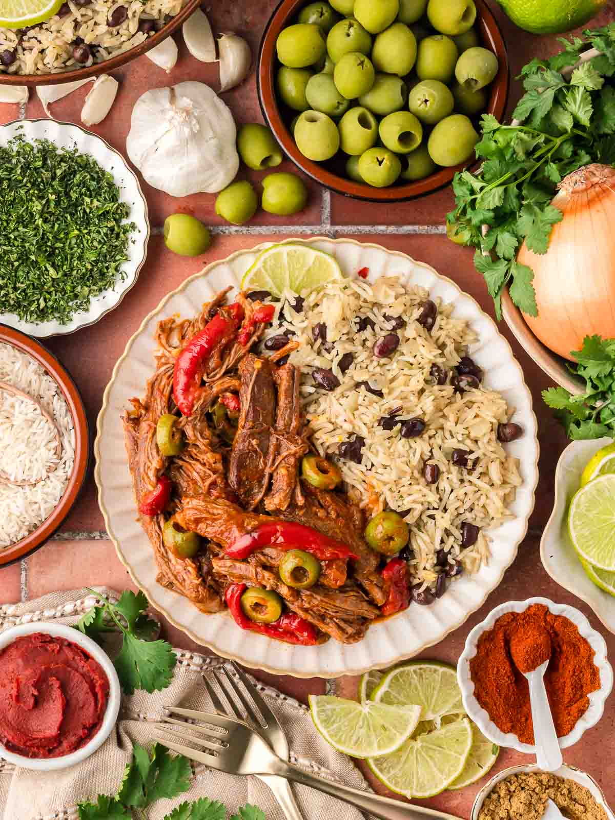 Plated slow cooked flank steak with red peppers and green olives served with rice and black beans, surrounded by fresh herbs, lime slices, and seasoning bowls.