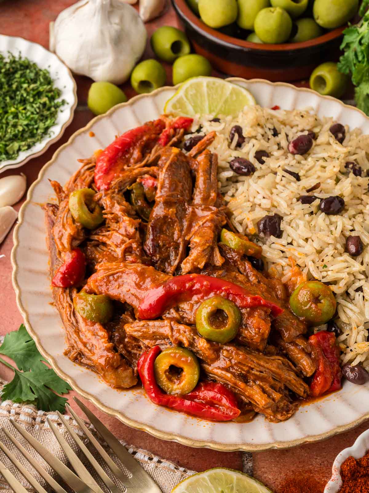 Close-up of shredded flank steak in tomato sauce with green olives and red peppers next to a serving of rice and black beans on a white scalloped plate.