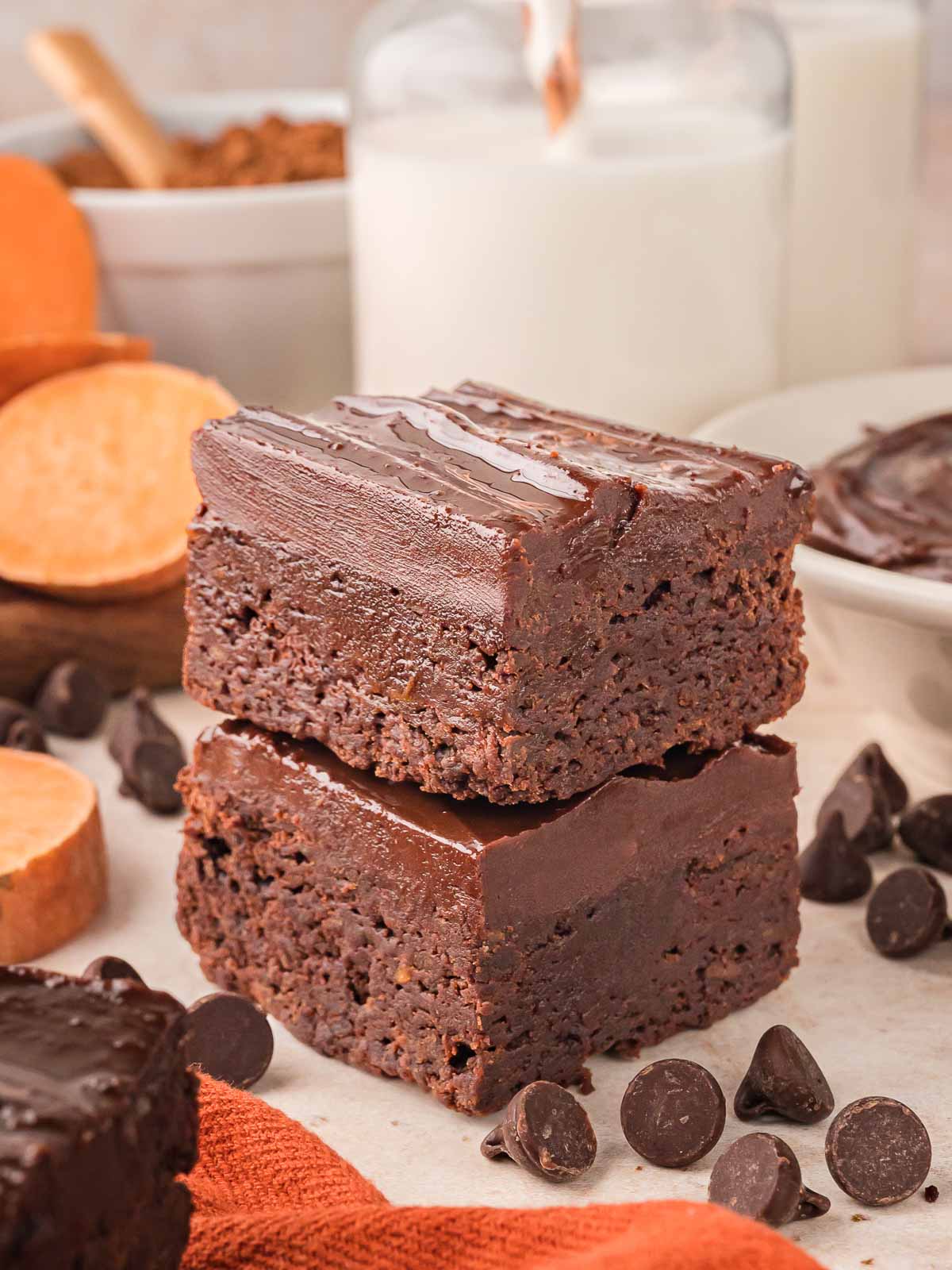Two thick sweet potato brownies stacked on top of each other, topped with smooth chocolate ganache, surrounded by chocolate chips and sweet potato slices.
