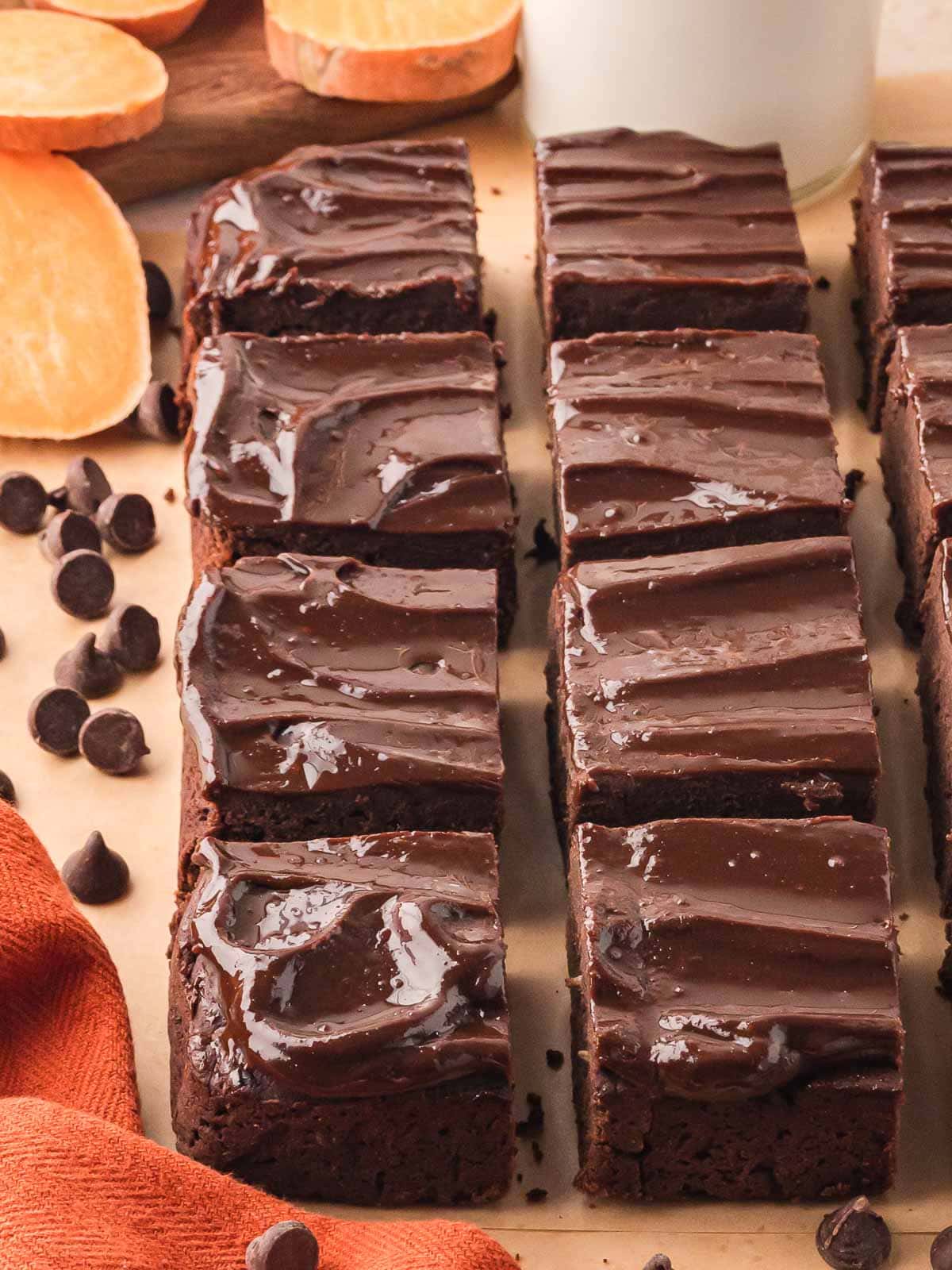 Rows of fudgy sweet potato brownies topped with glossy chocolate ganache, sliced into squares and arranged on parchment with sweet potato slices and chocolate chips.