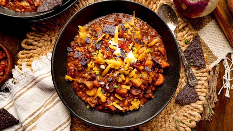 Bowl of pumpkin chili topped with sour cream, shredded cheddar, green onions, and crispy bacon, served with blue corn tortilla chips.