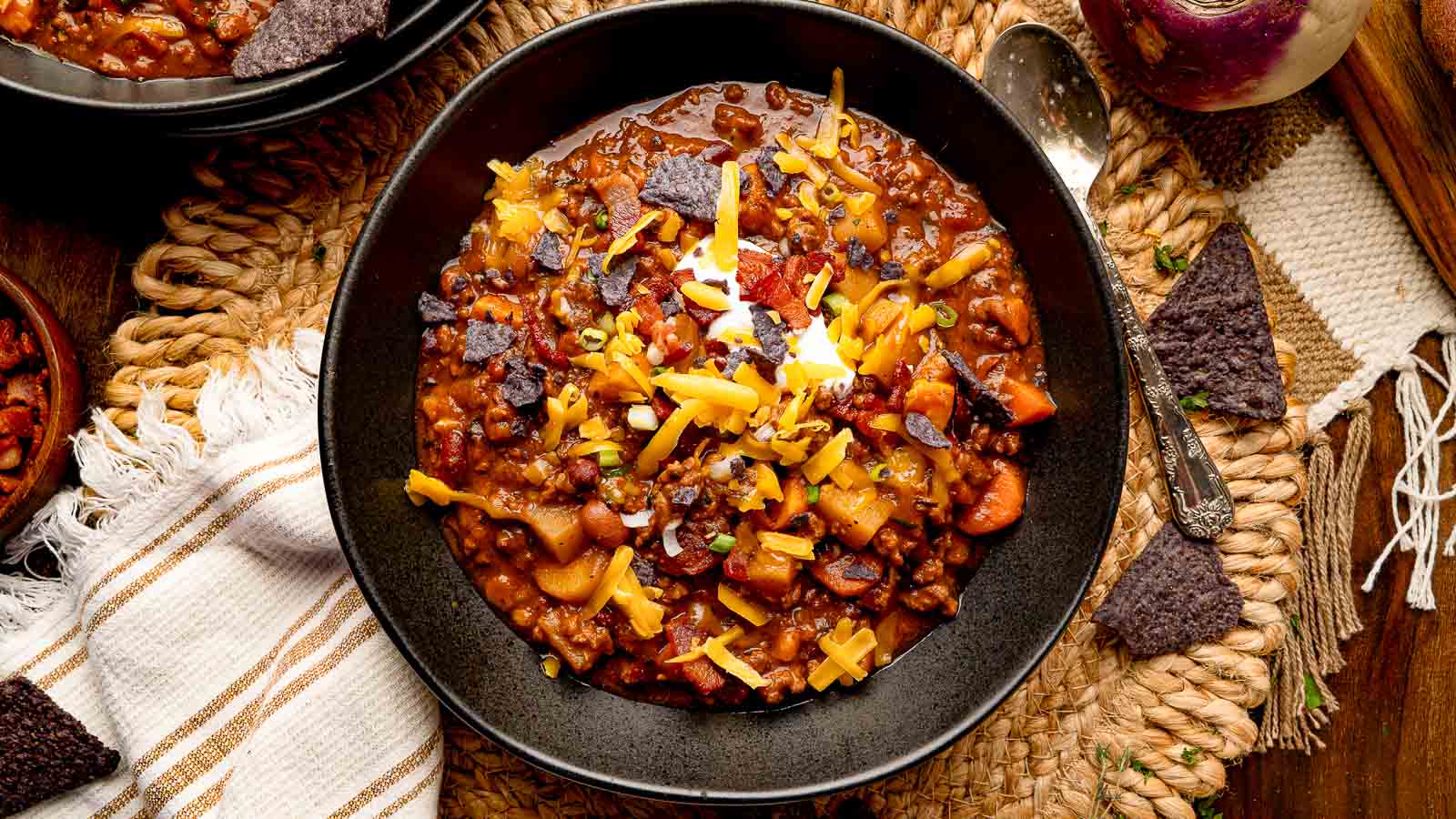 Bowl of pumpkin chili topped with sour cream, shredded cheddar, green onions, and crispy bacon, served with blue corn tortilla chips.