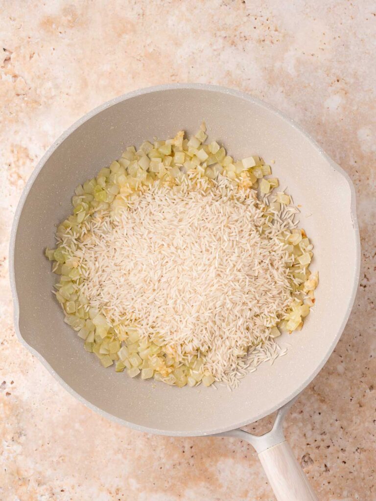 Uncooked white rice added to sautéed onions and garlic in the skillet, ready to be toasted.