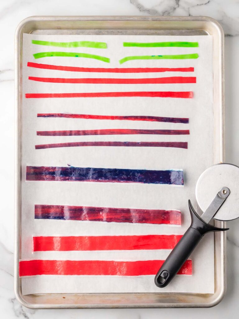 Strips of red, green, and purple fruit roll-ups cut on parchment paper with a pizza cutter on the side.
