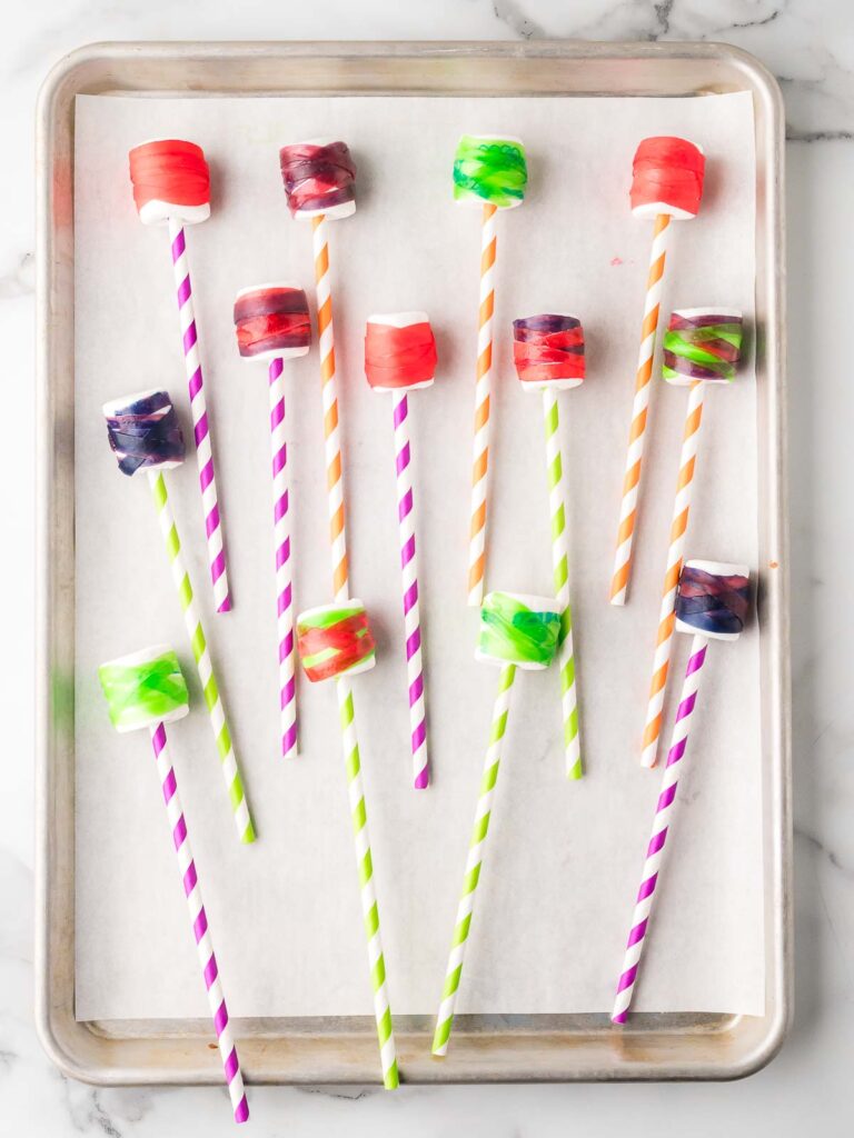 Marshmallows on colorful striped paper straws wrapped with fruit roll-up strips, arranged on a baking sheet.