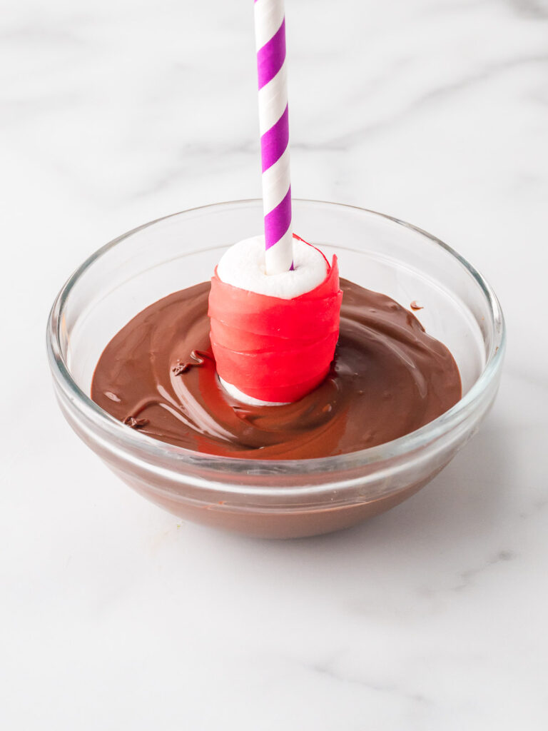 Marshmallow on a purple-striped straw wrapped in red fruit roll-up being dipped into melted chocolate.