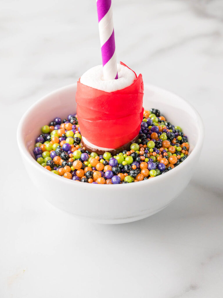 Marshmallow pop wrapped in red fruit roll-up being dipped into a bowl of Halloween-colored sprinkles.