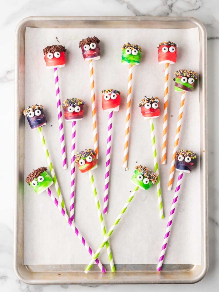 Decorated marshmallow monster pops with sprinkles and candy eyes on striped straws set on a parchment-lined baking sheet.
