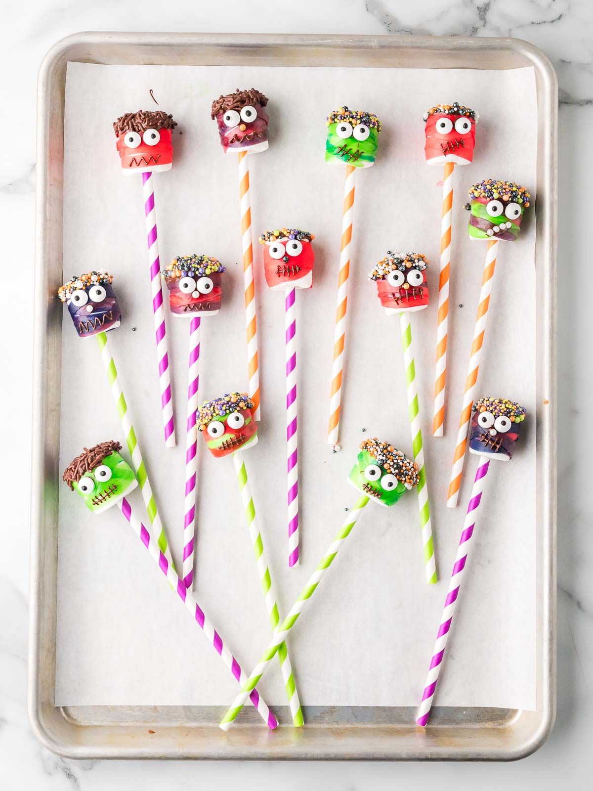 Tray of finished monster marshmallow pops decorated with candy eyeballs, sprinkles, and chocolate details on striped paper straws.