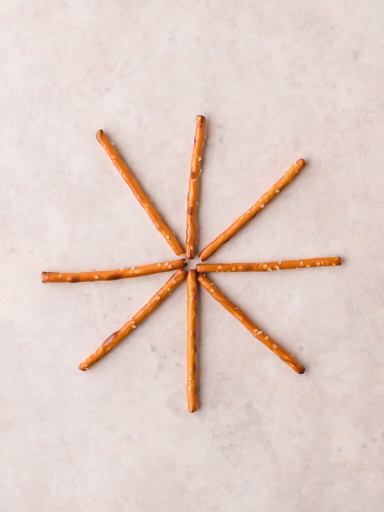 Eight pretzel sticks arranged in a starburst shape on parchment paper as the base for a Pretzel Spider Web.