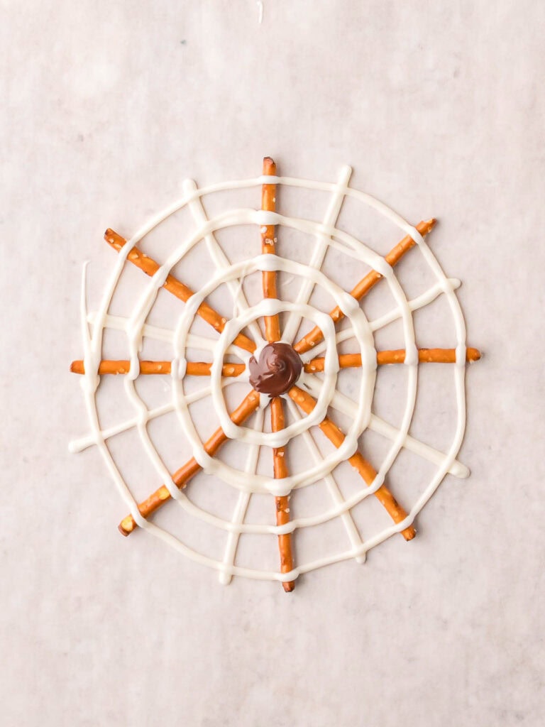 Pretzel Spider Web with white chocolate drizzle and milk chocolate center on parchment paper.