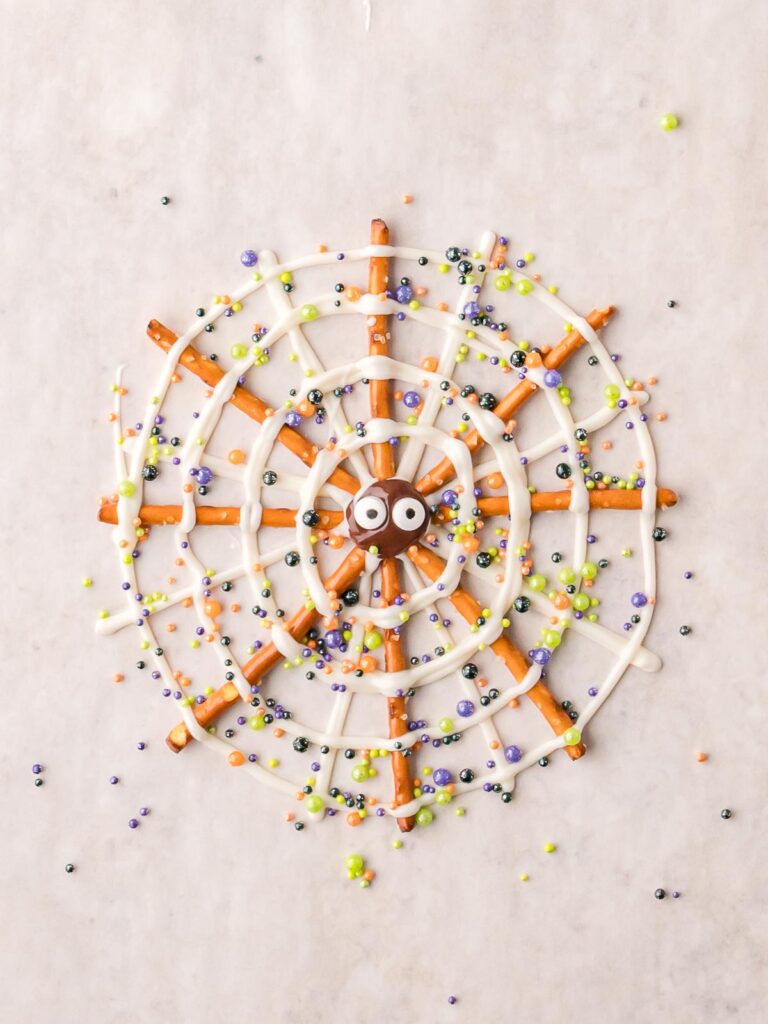 Pretzel Spider Web with white chocolate drizzle, candy eyes, and Halloween sprinkles on parchment paper.