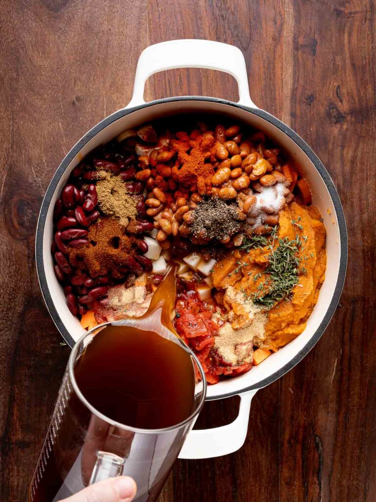 Beef broth being poured into a pot filled with chili ingredients before simmering.