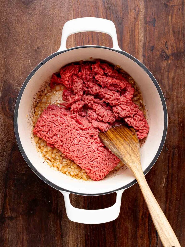 Raw ground beef added to the pot with sautéed onions and garlic, ready to be browned.