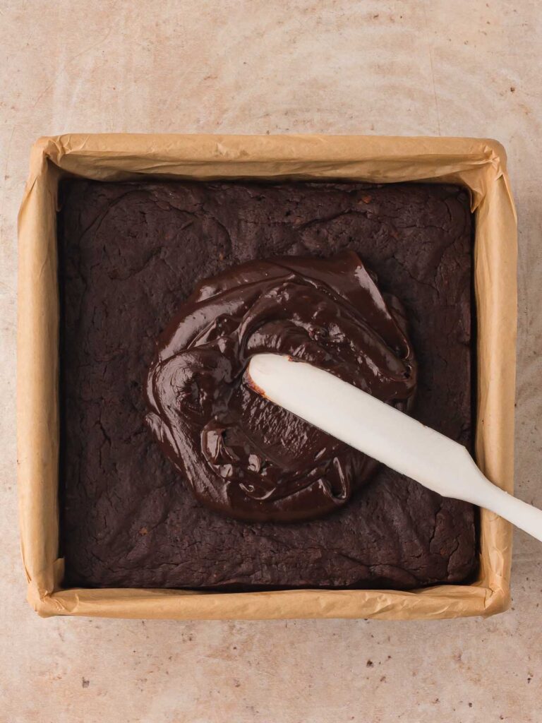Spreading chocolate ganache over the cooled sweet potato brownie base in a parchment-lined pan.