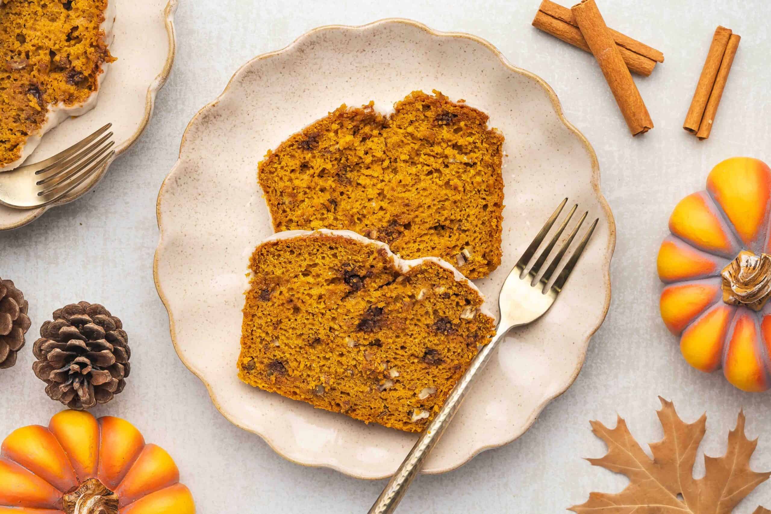 Two slices of moist pumpkin bread with cinnamon and nuts on a speckled cream plate, surrounded by pumpkins, pinecones, and cinnamon sticks.
