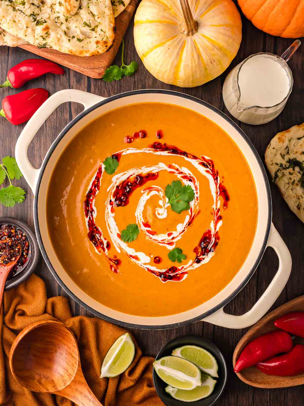 Finished pumpkin curry soup garnished with cream, chili oil, and cilantro, surrounded by naan bread, limes, and pumpkins.