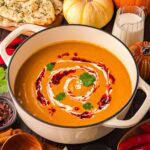 Creamy Thai-style pumpkin soup in a white Dutch oven with fall pumpkins, fresh naan, and chili oil around it.