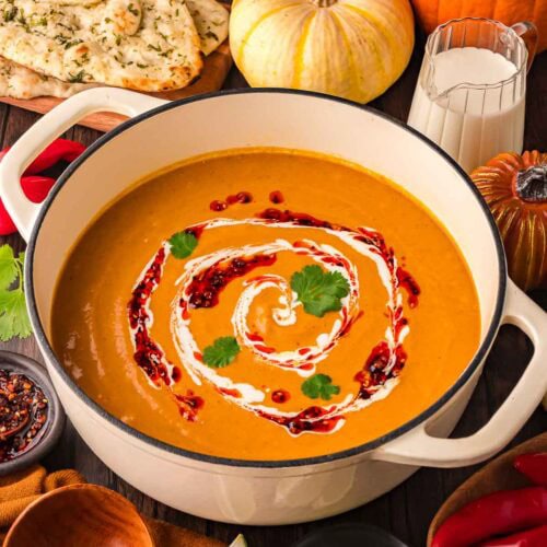 Creamy Thai-style pumpkin soup in a white Dutch oven with fall pumpkins, fresh naan, and chili oil around it.