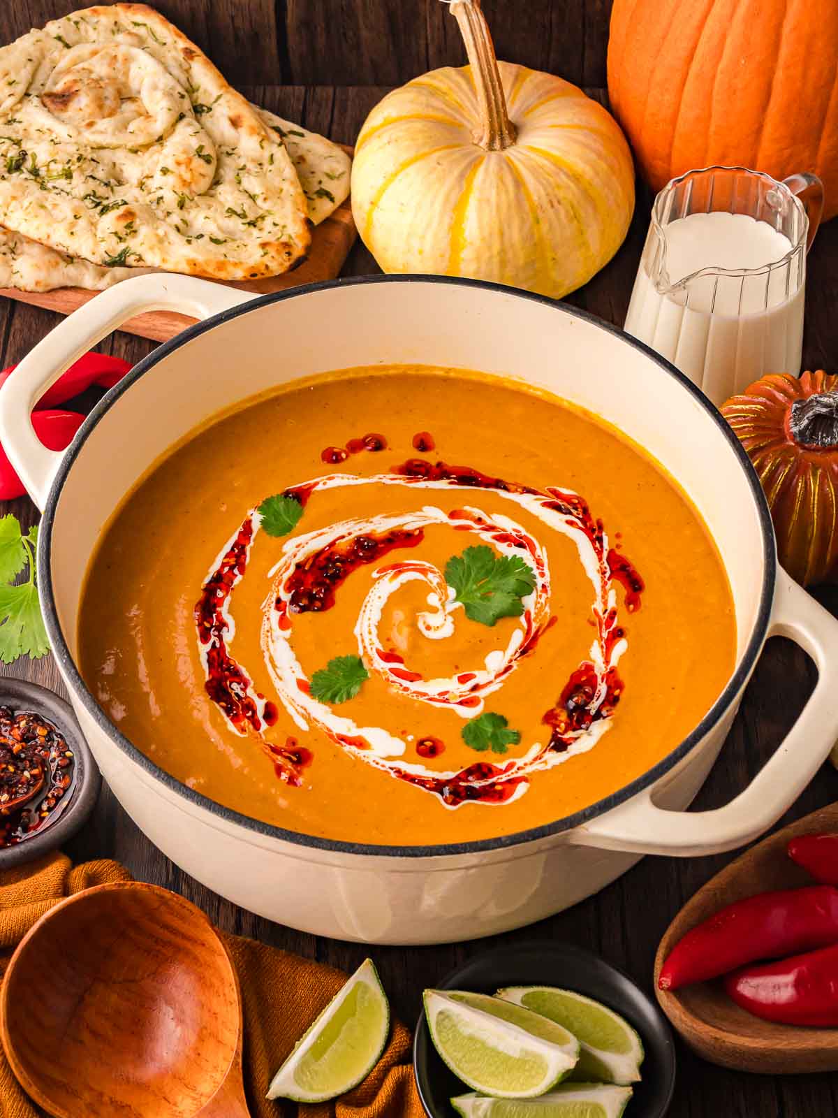Creamy Thai-style pumpkin soup in a white Dutch oven with fall pumpkins, fresh naan, and chili oil around it.