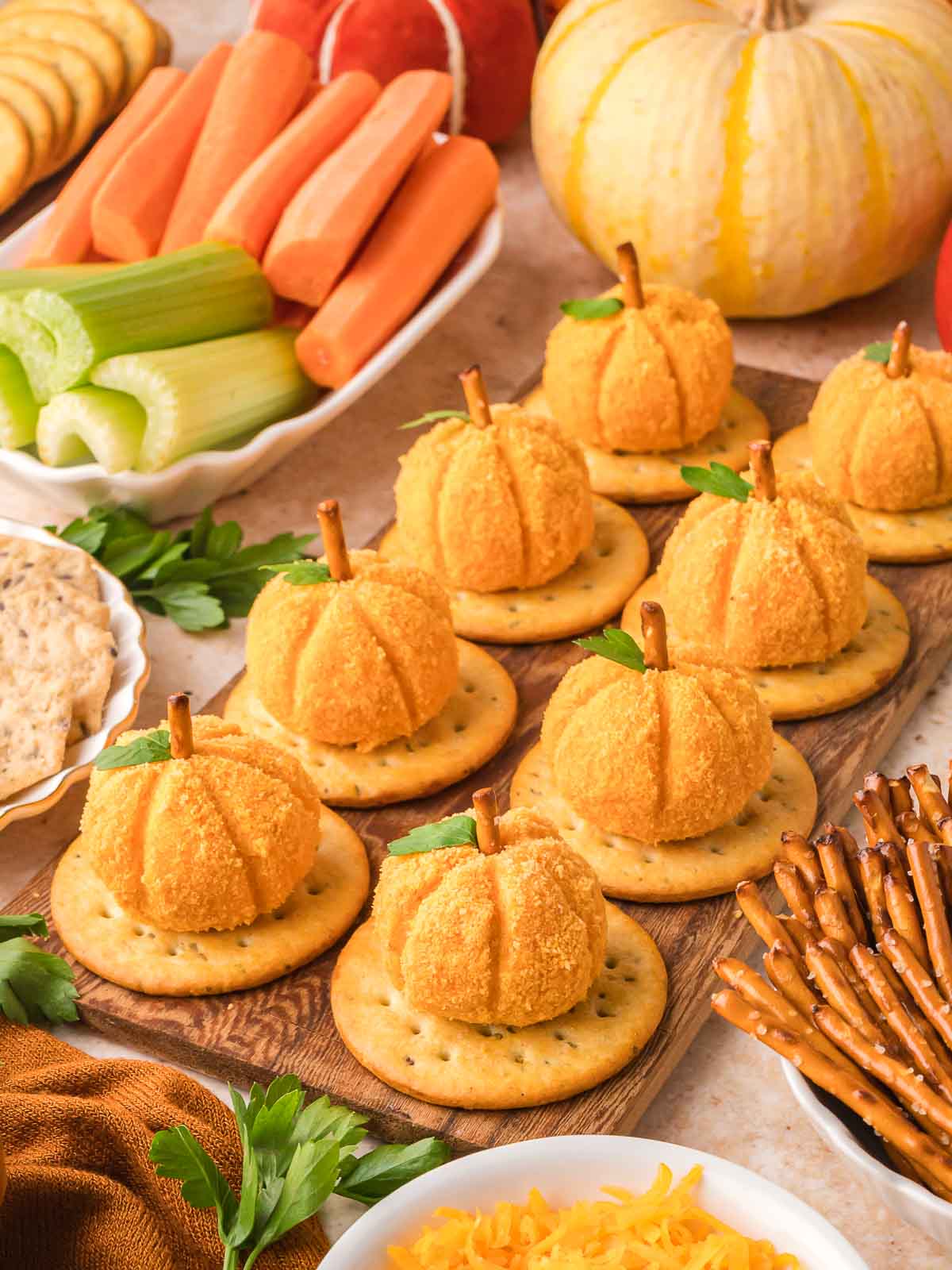 Mini pumpkin cheese balls on round crackers, garnished with parsley and pretzel stems, surrounded by fresh veggie sticks and white pumpkin decor.
