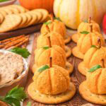 Side view of mini pumpkin-shaped cheese balls on crackers, arranged in a row with fall pumpkins and party crackers in the background.