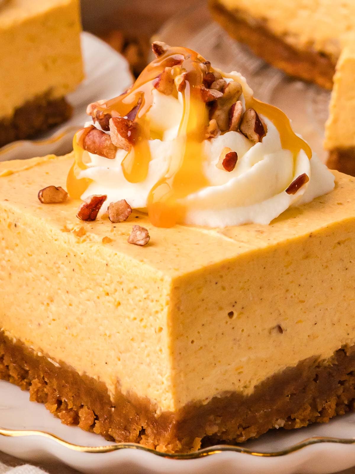 Slice of no-bake pumpkin cheesecake bar on a white plate, topped with whipped cream, chopped pecans, and caramel drizzle.