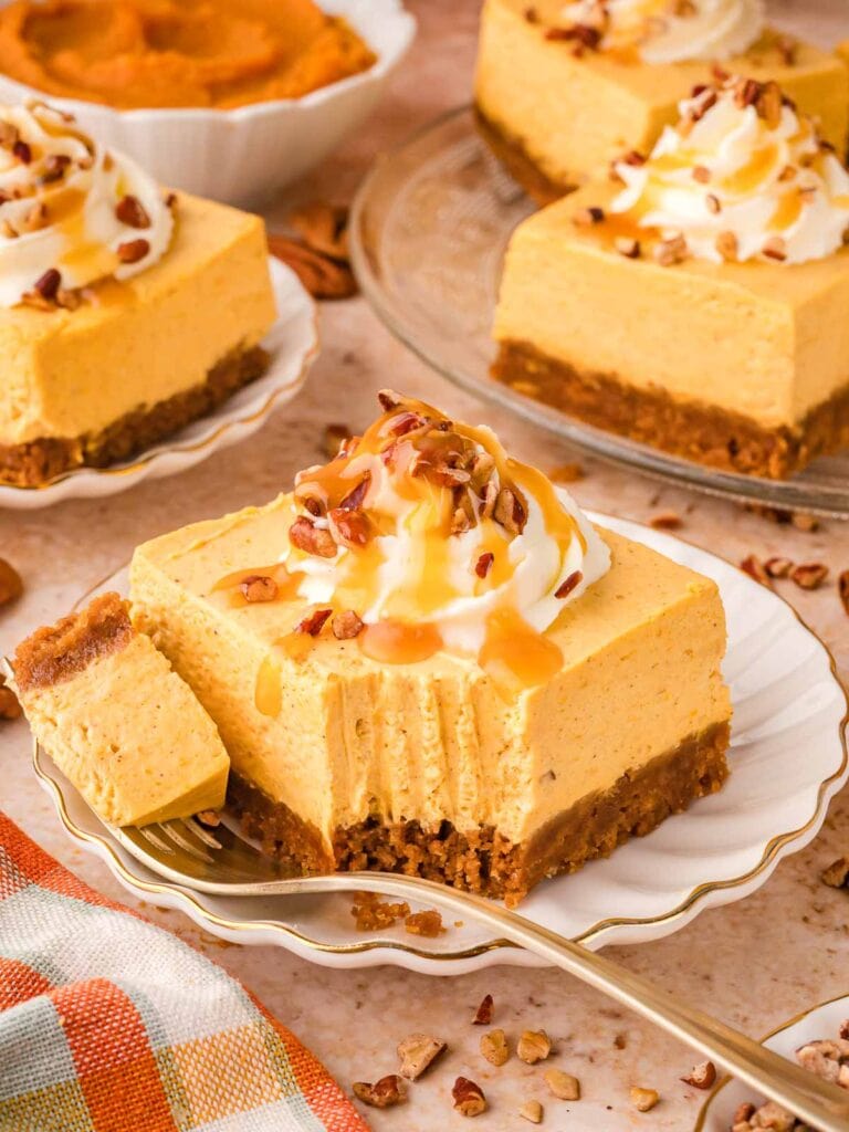 Plate of no-bake pumpkin cheesecake bar with a bite taken out, surrounded by more bars and bowls of pumpkin purée and pecans.