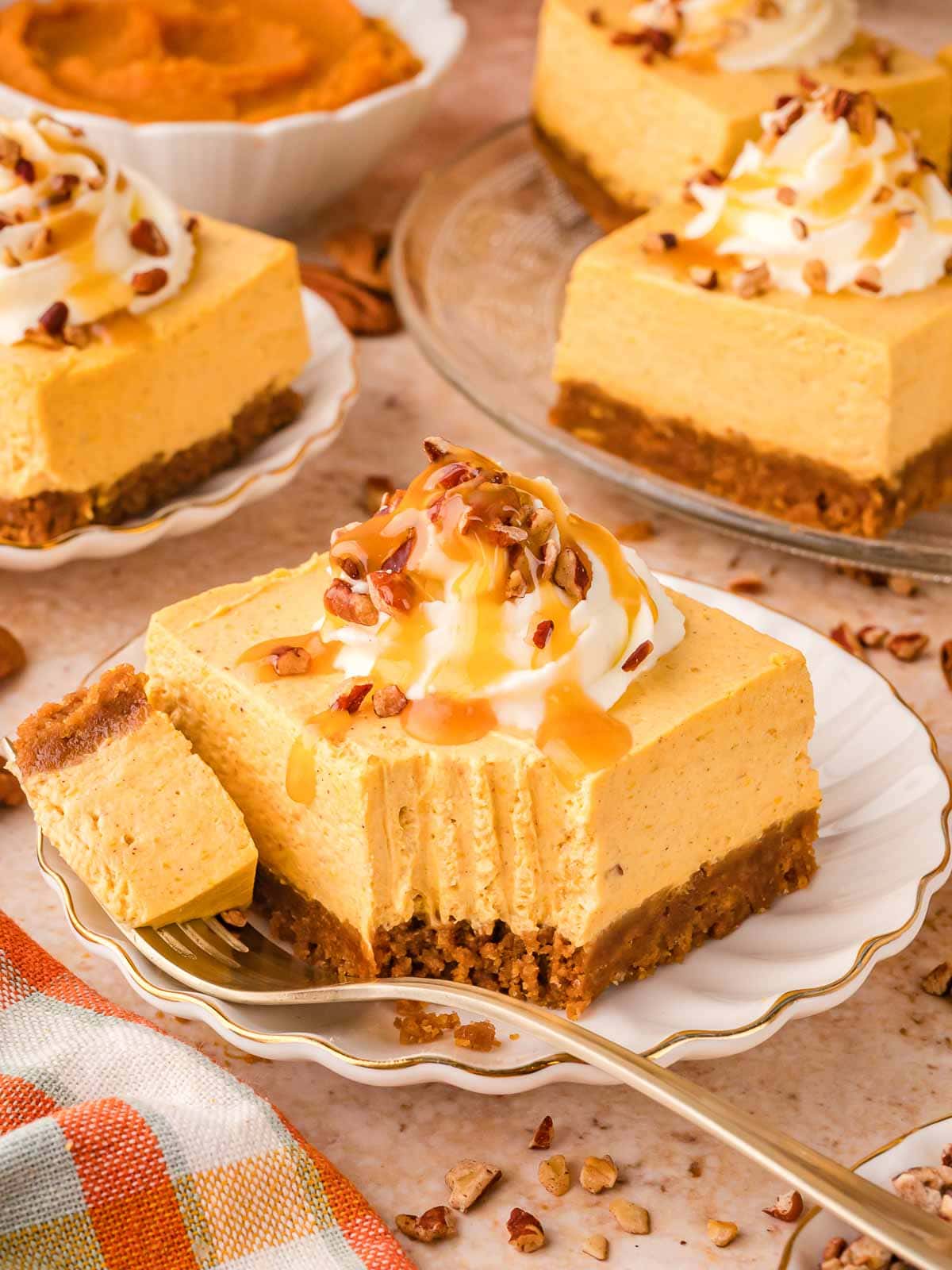 Plate of no-bake pumpkin cheesecake bar with a bite taken out, surrounded by more bars and bowls of pumpkin purée and pecans.