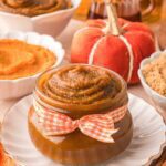 Pumpkin butter in a small glass jar tied with an orange gingham ribbon, styled with mini pumpkins, maple syrup, and brown sugar.