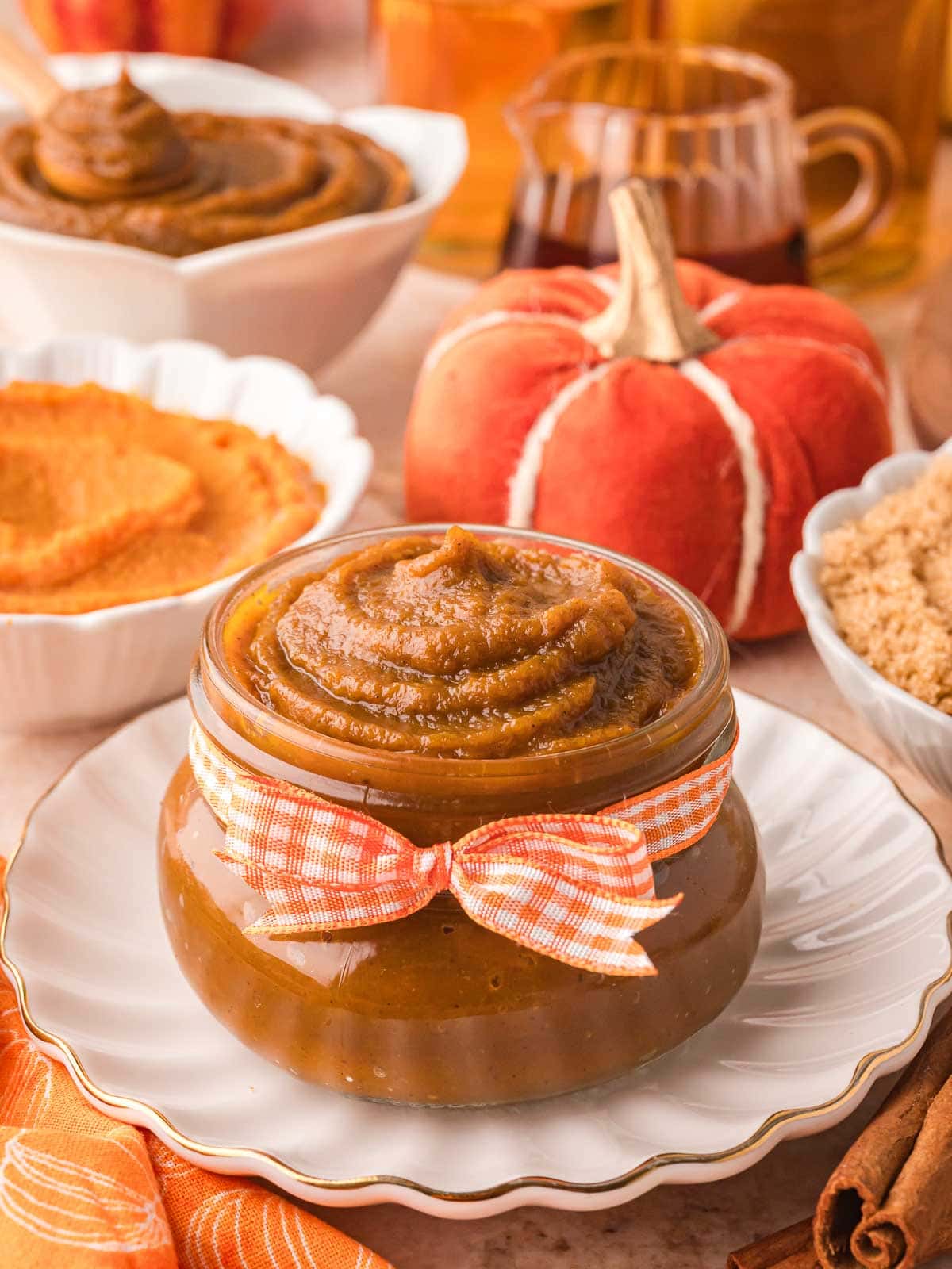 Pumpkin butter in a small glass jar tied with an orange gingham ribbon, styled with mini pumpkins, maple syrup, and brown sugar.