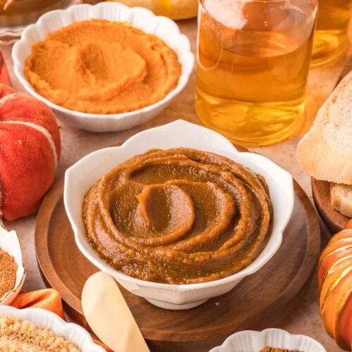 Bowl of thick homemade pumpkin butter surrounded by canned pumpkin, apple juice, brown sugar, and fall spices on a wooden surface.