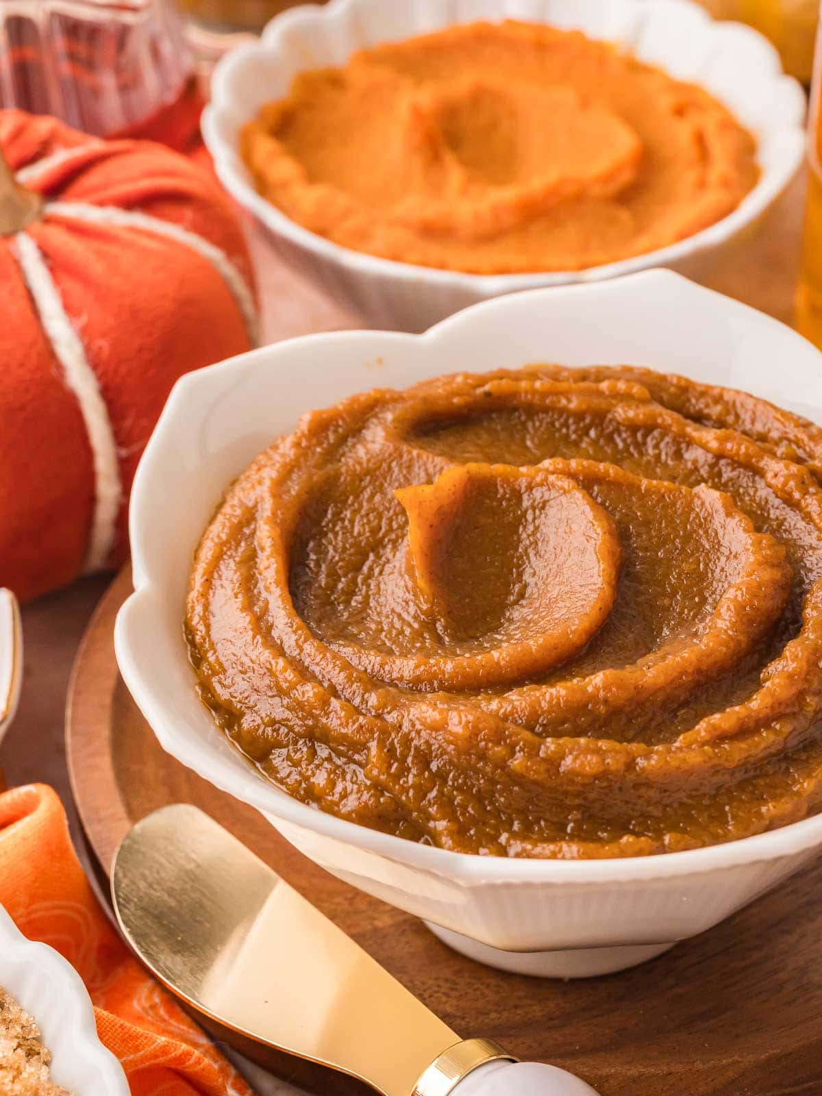 Smooth and glossy pumpkin butter swirled in a white bowl, with a gold spreader and fall-themed ingredients in the background.