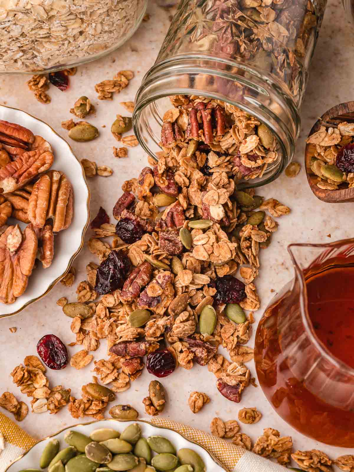 Pumpkin granola poured from a jar surrounded by pecans, pumpkin seeds, and a pitcher of maple syrup.
