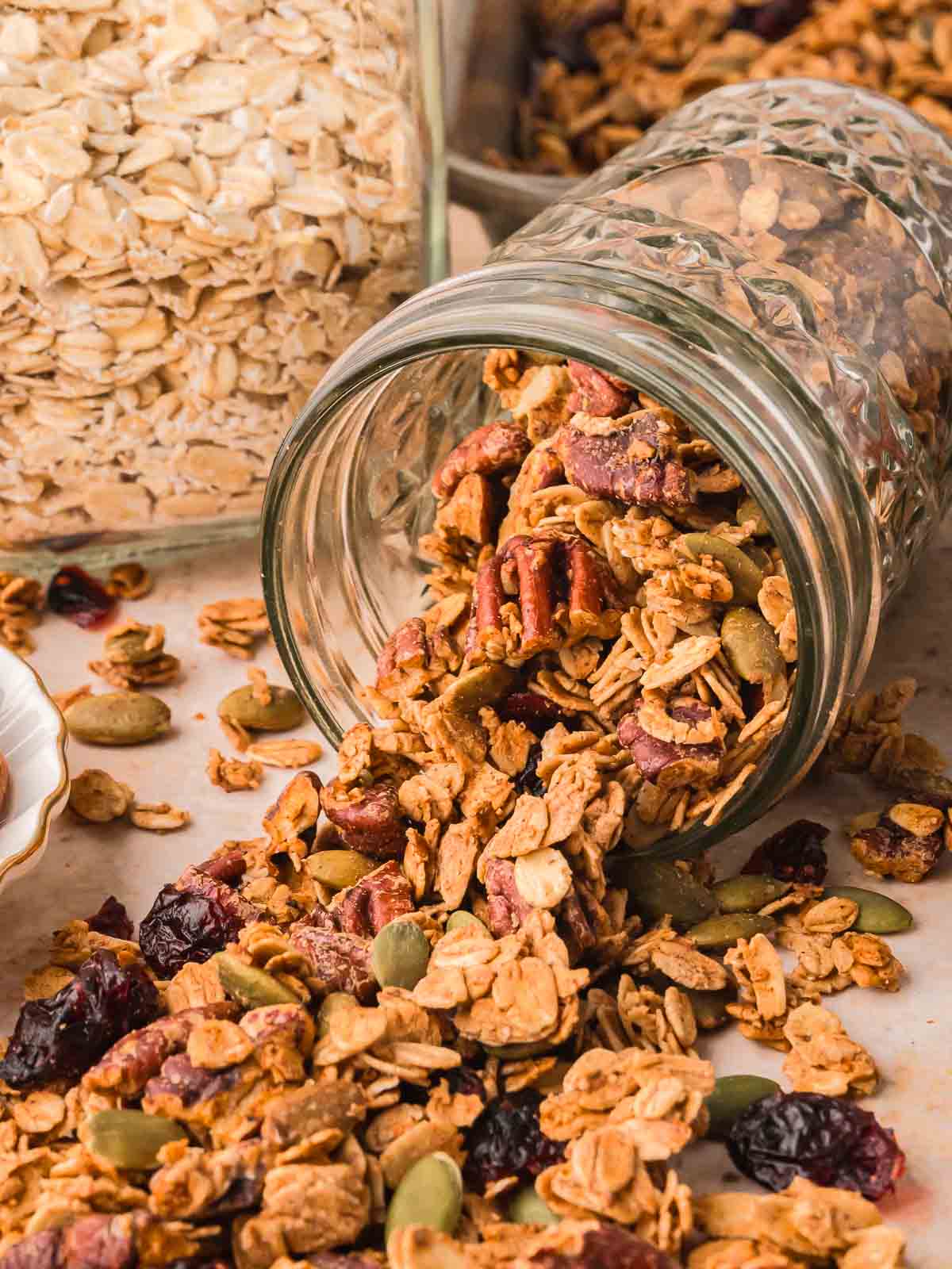 Pumpkin granola spilling from a glass jar with oats, nuts, and cranberries scattered on the table.