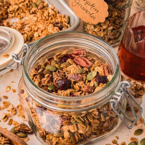 Jar of pumpkin granola with ribbon-tied gift jar and tray of baked granola in the background.
