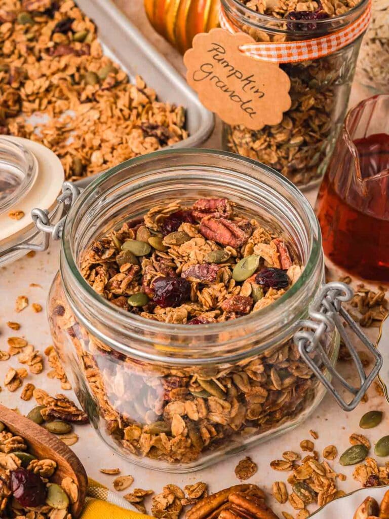 Jar of pumpkin granola with ribbon-tied gift jar and tray of baked granola in the background.