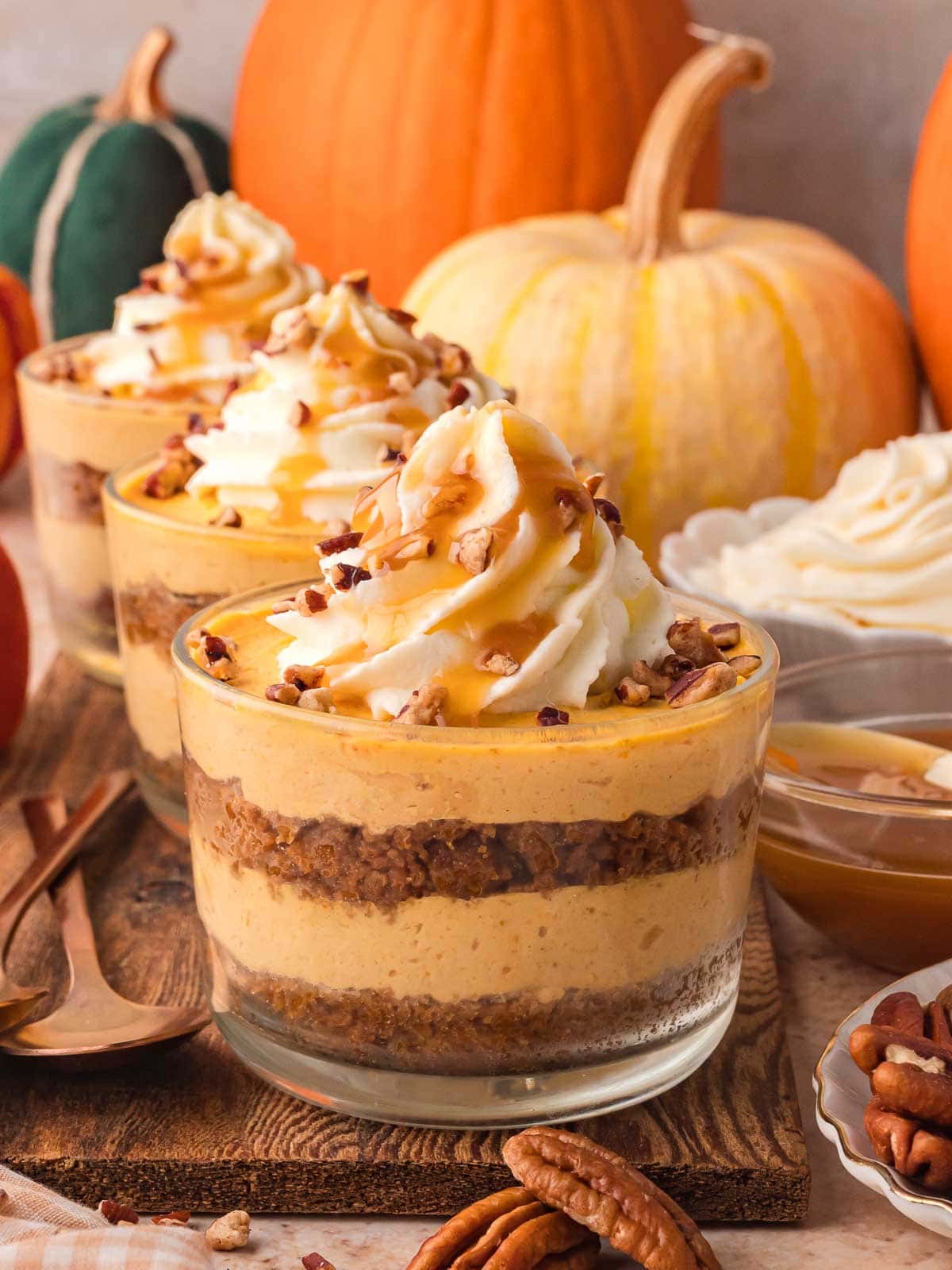 Three pumpkin pie cheesecake cups on a wooden board, showing layers of Biscoff crust and pumpkin cheesecake filling, topped with whipped cream, pecans, and caramel.