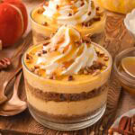 Two pumpkin pie cheesecake cups layered with Biscoff crust and pumpkin filling, topped with whipped cream and caramel, styled with pecans and pumpkins.