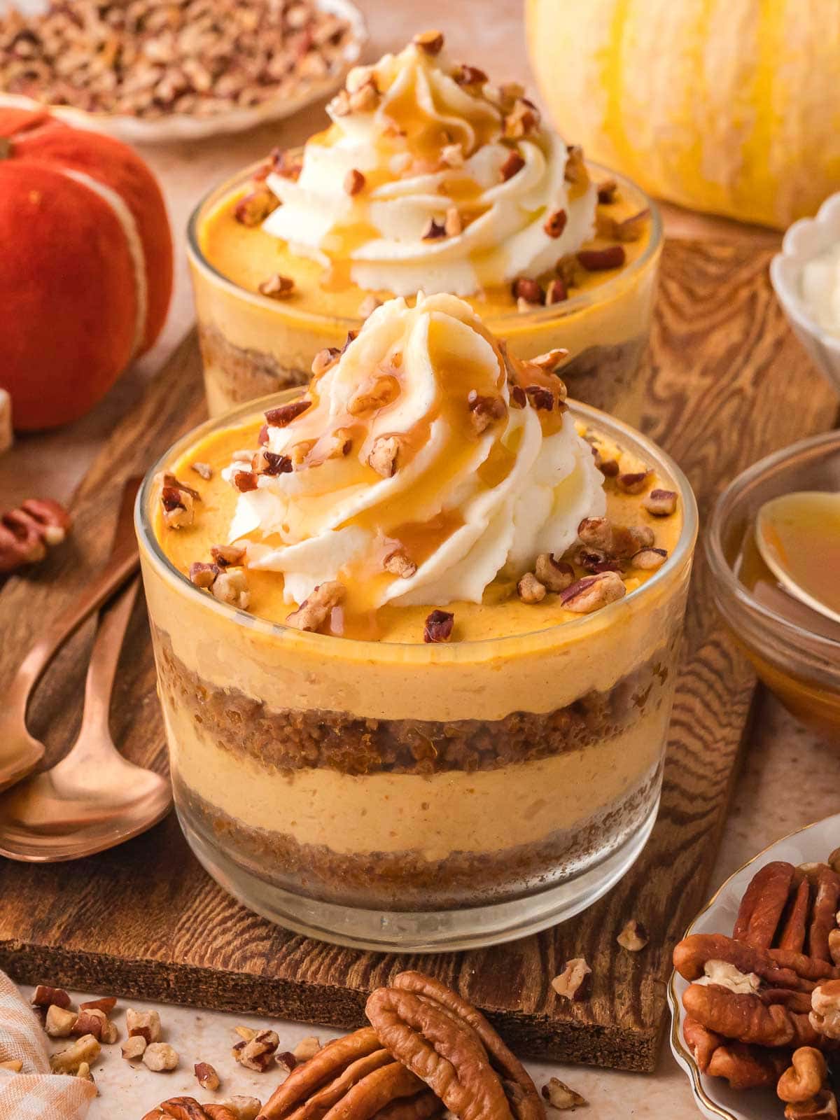 Two pumpkin pie cheesecake cups layered with Biscoff crust and pumpkin filling, topped with whipped cream and caramel, styled with pecans and pumpkins.