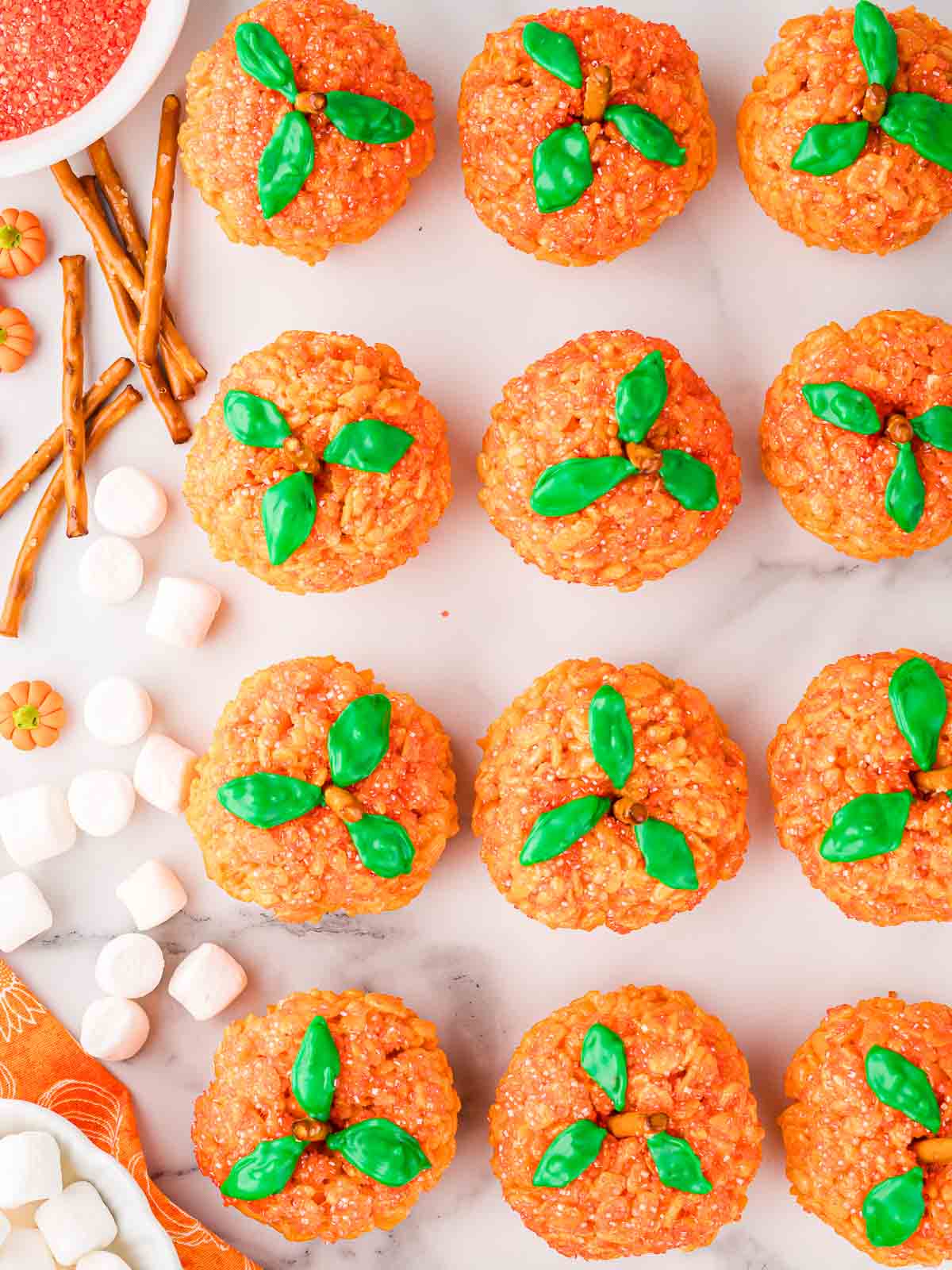 Flat lay of decorated Rice Krispie Pumpkins with green icing leaves and pretzel stalks, surrounded by marshmallows and pretzel sticks.