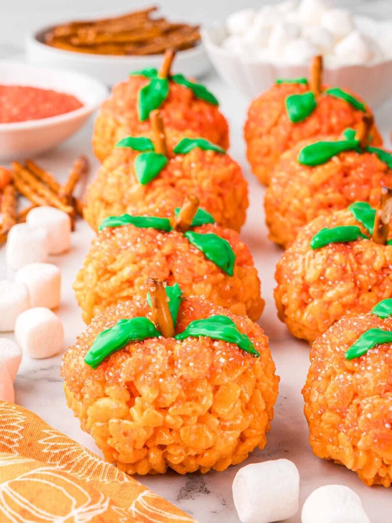 Front view of multiple Rice Krispie Pumpkins on a table with decorating ingredients in the background.