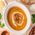 Slow cooker pumpkin soup served in a white crock with swirls of cream, crispy bacon, and chopped parsley, surrounded by sliced bread, parmesan, and fresh herbs.