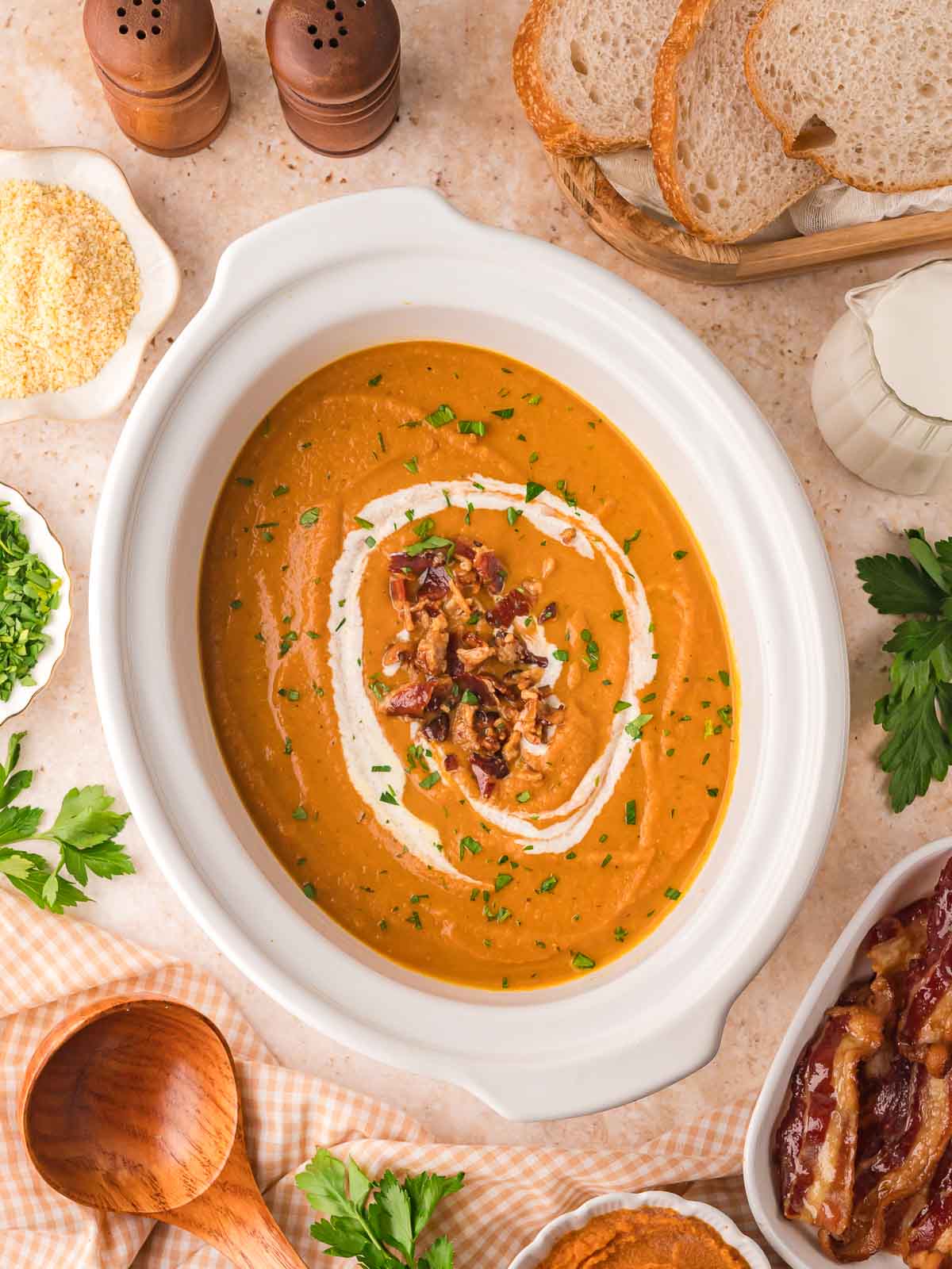 Slow cooker pumpkin soup served in a white crock with swirls of cream, crispy bacon, and chopped parsley, surrounded by sliced bread, parmesan, and fresh herbs.