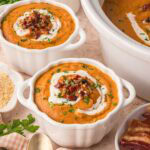 Two individual white bowls of slow cooker pumpkin soup with cream swirls, chopped bacon, and parsley, set beside the crockpot.