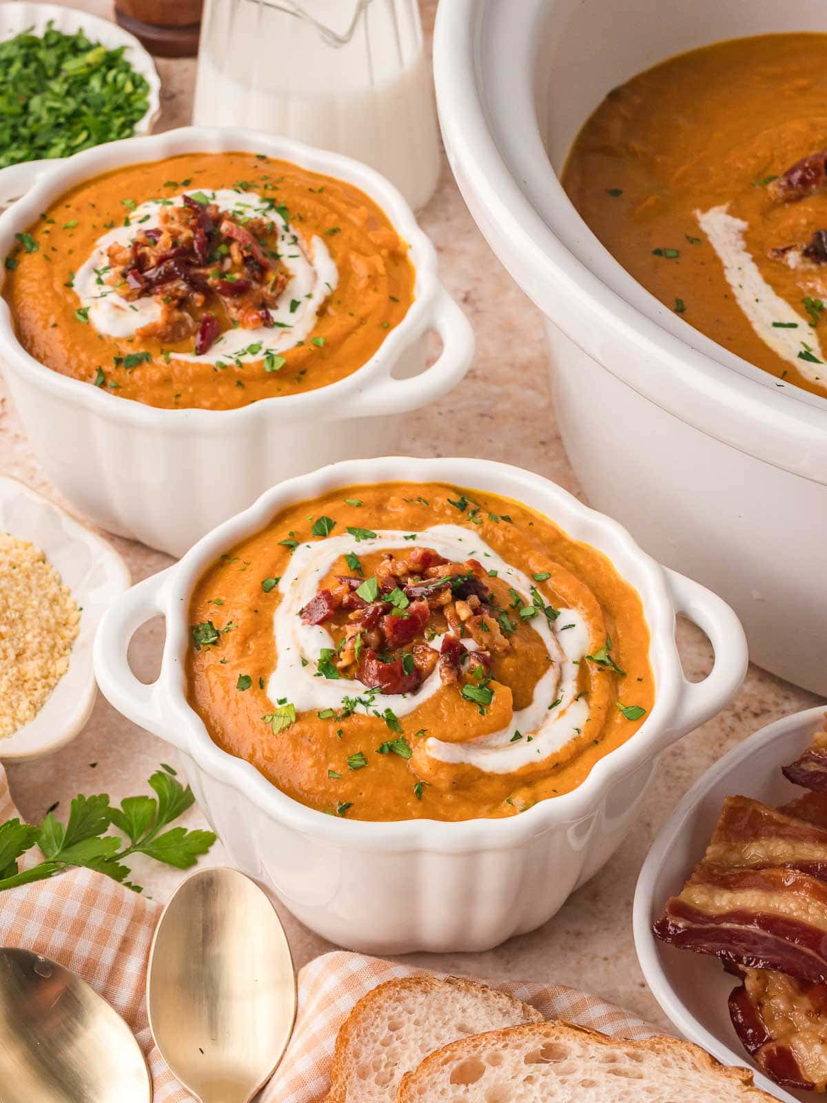 Two individual white bowls of slow cooker pumpkin soup with cream swirls, chopped bacon, and parsley, set beside the crockpot.