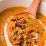 Wooden spoon lifting creamy slow cooker pumpkin soup topped with crispy bacon and herbs from a white crockpot.