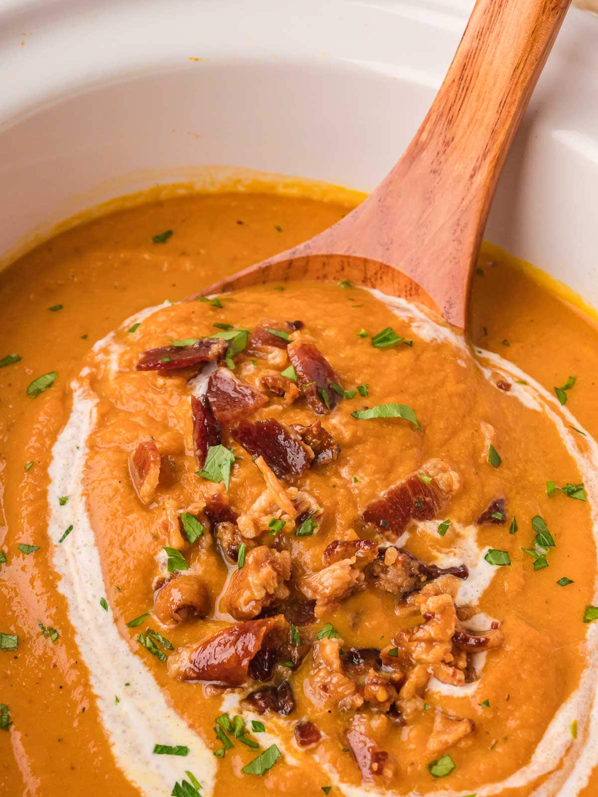 Wooden spoon lifting creamy slow cooker pumpkin soup topped with crispy bacon and herbs from a white crockpot.