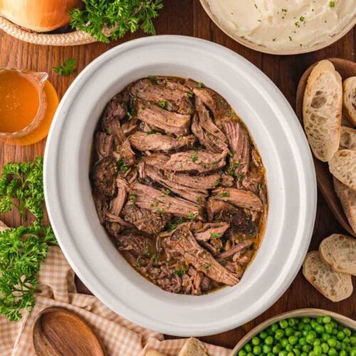 Slow cooker shredded beef served with mashed potatoes, peas, crusty bread, and fresh parsley.