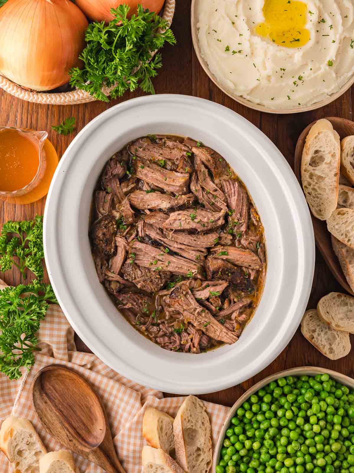 Slow cooker shredded beef served with mashed potatoes, peas, crusty bread, and fresh parsley.